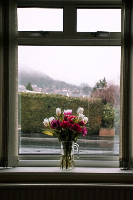 A clear glass vase filled with a fresh floral arrangement featuring white tulips and vibrant pink carnations, positioned on a windowsill. The bouquet is wrapped loosely at the base, showcasing healthy