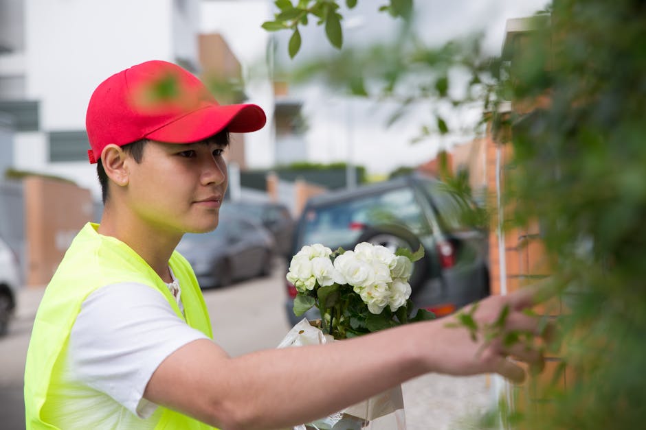 A young delivery person wearing a red cap and a high-visibility yellow vest is carefully handling a floral gift, consisting of fresh white hydrangeas, as they approach a house on a residential street.