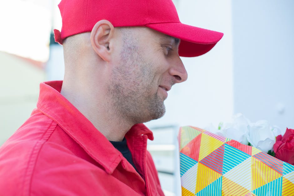 A delivery person dressed in a red uniform with a matching red cap is holding a colorful floral gift box wrapped in geometric patterned paper with shades of yellow, blue, pink, and white. Inside the b