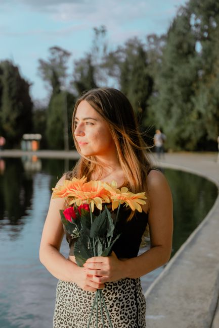 A young woman with light brown hair, styled naturally and softly flowing in the breeze, is holding a bouquet of bright, fresh flowers near a tranquil lake at Highams Park. The bouquet features large,