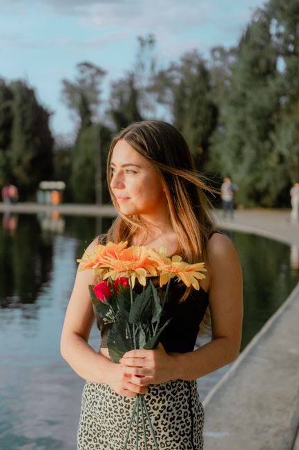 A young woman with long brown hair, wearing a black sleeveless top and leopard print skirt, is standing by a lakeside path holding a bouquet of fresh flowers. The arrangement features vibrant peach an