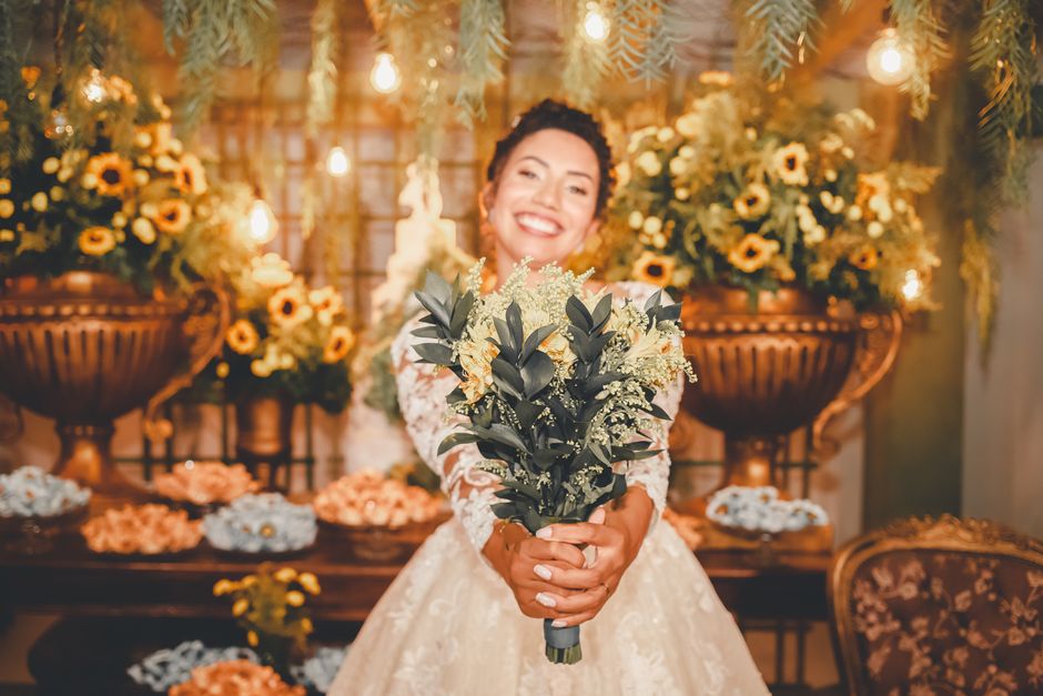 A smiling woman in a white lace wedding dress holds a bouquet of fresh flowers featuring dark green foliage and light cream blossoms. She stands in a warmly lit room decorated with abundant floral arr