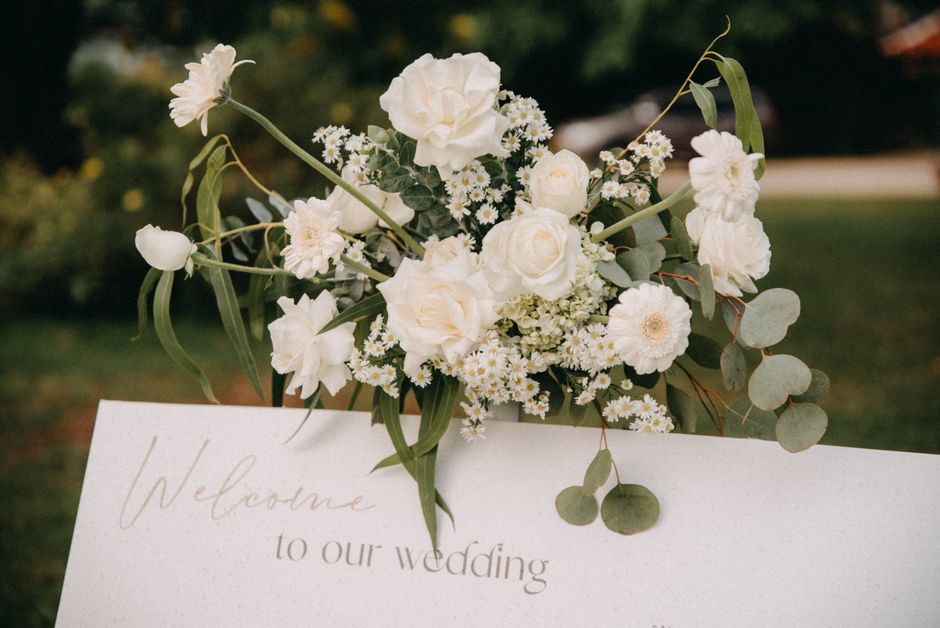 A delicate floral arrangement featuring white roses, small white daisy-like flowers, and light green eucalyptus leaves, arranged atop a white welcome sign with elegant cursive text that reads 'Welcome