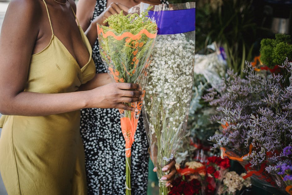 A person wearing a yellow satin dress holds a bouquet of mixed flowers wrapped in clear cellophane with orange accents. The bouquet features green and purple blooms, possibly including sedum and delic