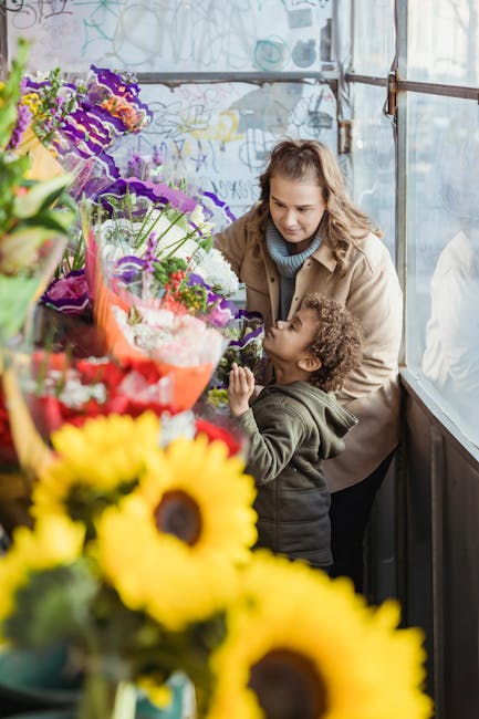 A young woman with wavy brown hair, wearing a light blue sweater and beige coat, is smiling while helping a small child with curly hair look at a vibrant display of fresh flowers in a greenhouse or fl
